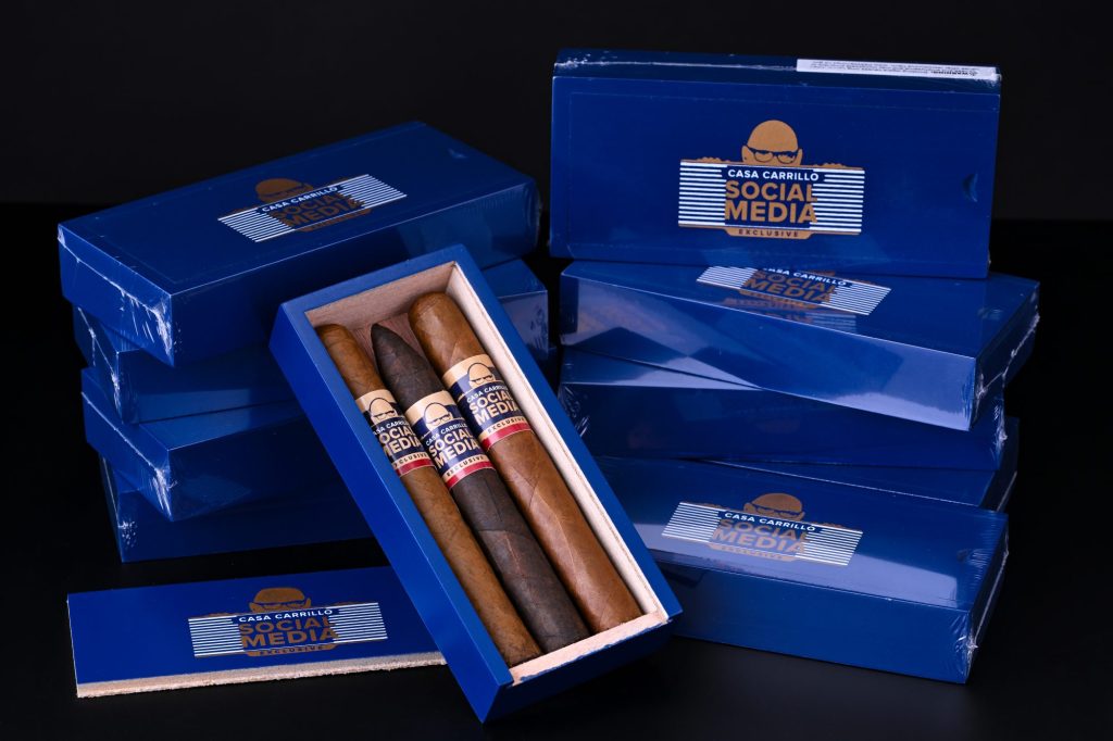 Casa Carrillo Social Media Exclusive cigar boxes stacked and displayed with one open box showing three cigars inside, featuring blue packaging and branded bands against a dark background.