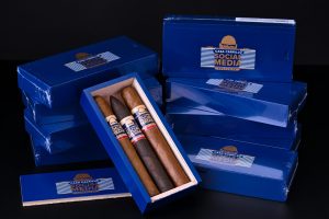 Casa Carrillo Social Media Exclusive cigar boxes stacked and displayed with one open box showing three cigars inside, featuring blue packaging and branded bands against a dark background.