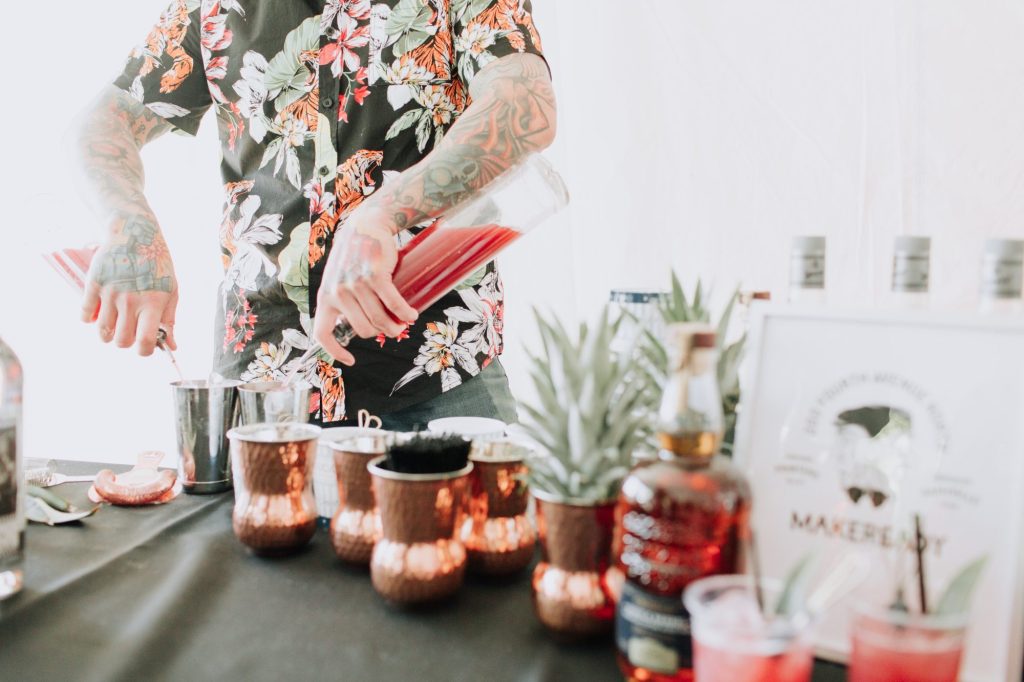 Bartender with tattooed arms pouring a red cocktail into copper mugs at an event table, surrounded by bar tools, fresh garnishes, and bottled spirits in a bright indoor setting.