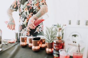 Bartender with tattooed arms pouring a red cocktail into copper mugs at an event table, surrounded by bar tools, fresh garnishes, and bottled spirits in a bright indoor setting.
