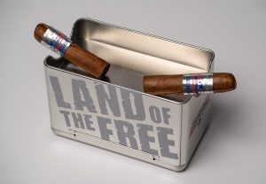 Two Independence Firecracker cigars resting inside an open metal cigar tin labeled “Land of the Free” on a white background.