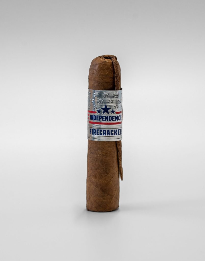 Independence Firecracker cigar standing upright on a white background, featuring a silver band with stars and red, white, and blue patriotic design.