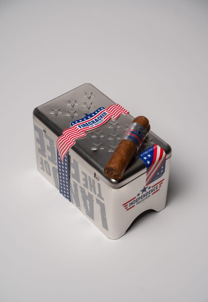 Independence Premium Cigars Firecracker cigar resting on a patriotic metal cigar tin featuring stars and an American flag design.