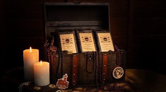 Open wooden chest displaying boxed cigars with pirate-themed labels, surrounded by candles, coins, and bead necklaces in a dark, atmospheric setting.