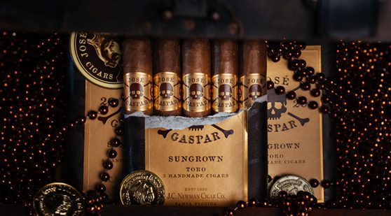 Open package of J.C. Newman José Gaspar Sungrown Toro cigars displayed with skull-and-crossbones branding, coins, and bead necklaces on a dark background.