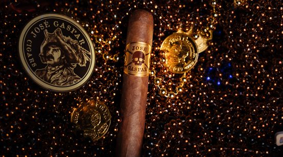 osé Gaspar cigar displayed on a bed of dark beads with gold coins and a round José Gaspar Cigars emblem in a dramatic, low-light setting.