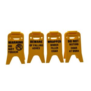 Bright yellow 3D printed cigar holder styled like a classic wet floor caution sign, printed with humorous cigar-related phrases in black text. The foldable design allows the sign to stand upright and serve as a portable cigar rest that keeps cigars elevated and off tables.