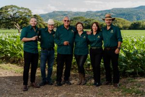 6 people standing side by side smoking cigars. In the background is a tobacco field.